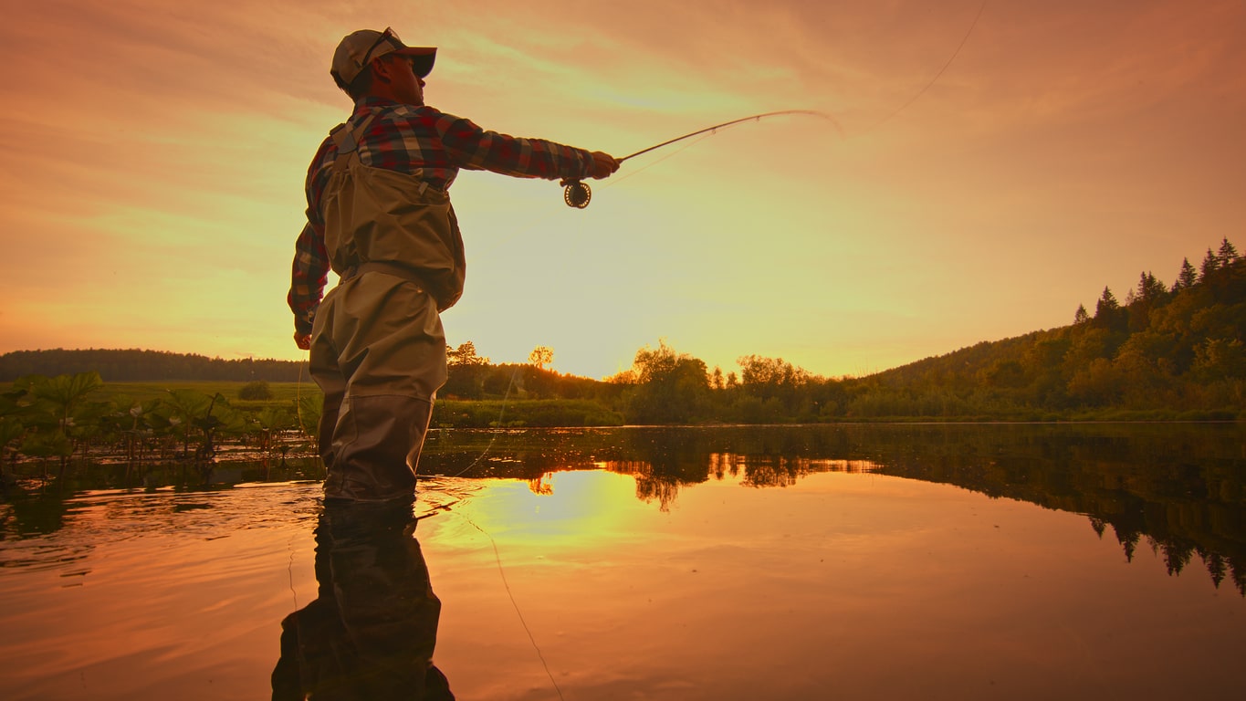 Fly fisherman stands in the water fishing on the river at sunset