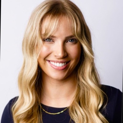 Smiling woman with long blonde hair and bangs, wearing a black top and gold necklace, on a light background.