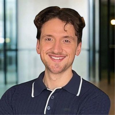 Smiling man with short brown hair wearing a dark polo shirt, standing indoors with glass background.