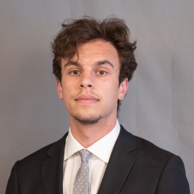 Young man with wavy brown hair in a suit and tie, posing against a plain gray background.